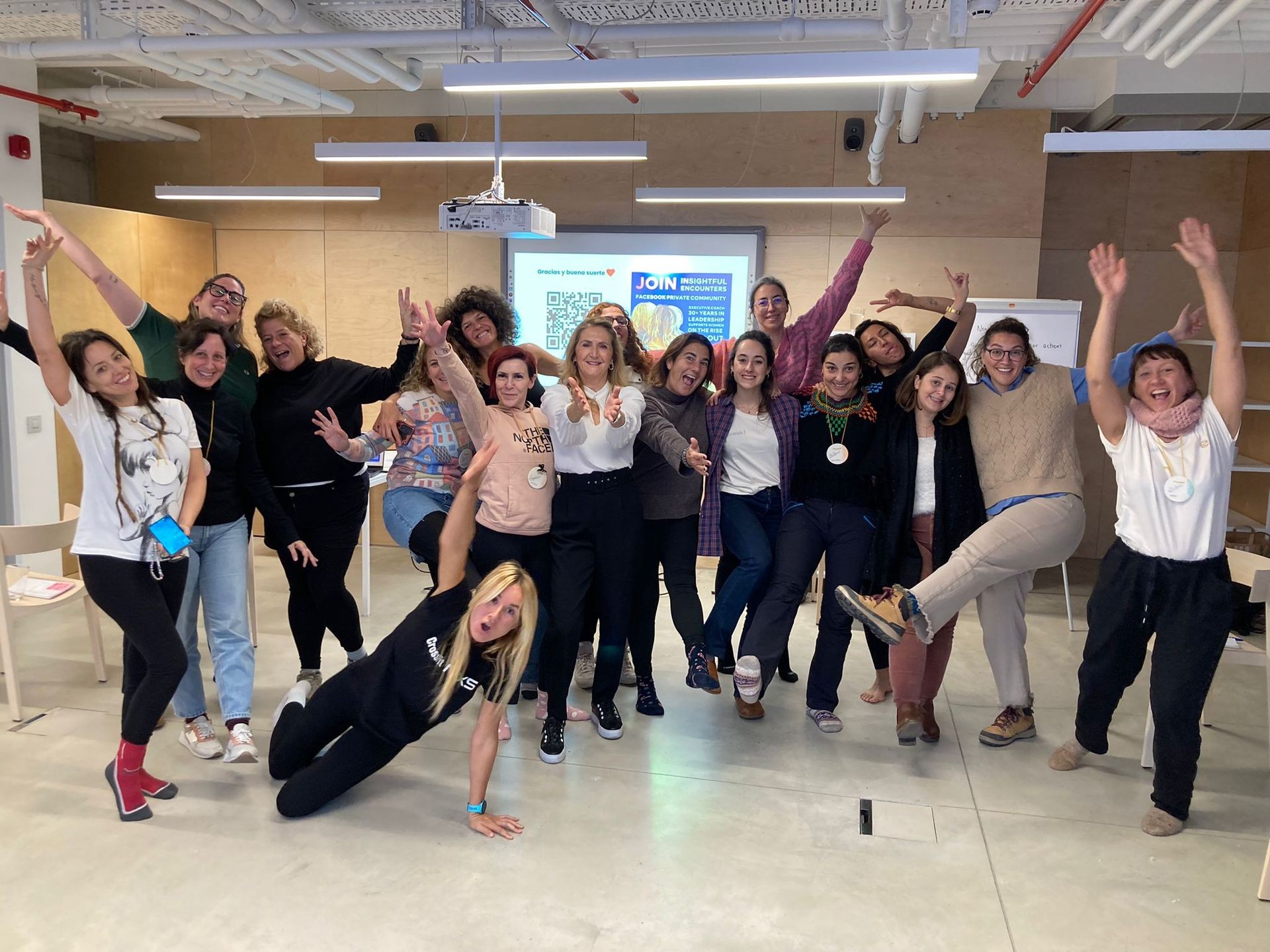 A cheerful group of women standing together in an indoor space, smiling, laughing, and posing with energetic hand gestures. The setting includes modern office lighting, a beige wall, and a visible projector on the ceiling. A cheerful group of women standing together in an indoor space, smiling, laughing, and posing with energetic hand gestures. The setting includes modern office lighting, a beige wall, and a visible projector on the ceiling.